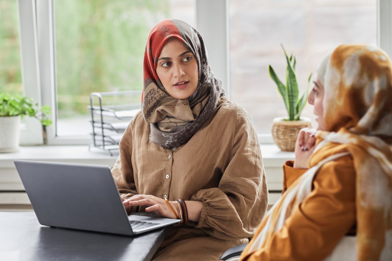 Home Two women in hijabs work together in a modern office, discussing at a laptop.